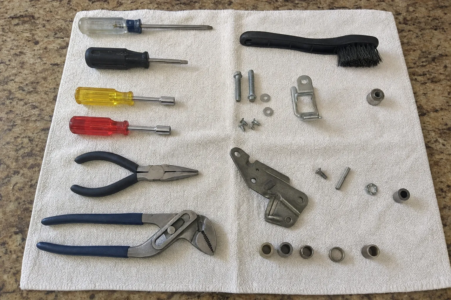 Screwdrivers, nut drivers and pliers on a towel beside a removed hinge arm, bracket, bolts, washers and bushings